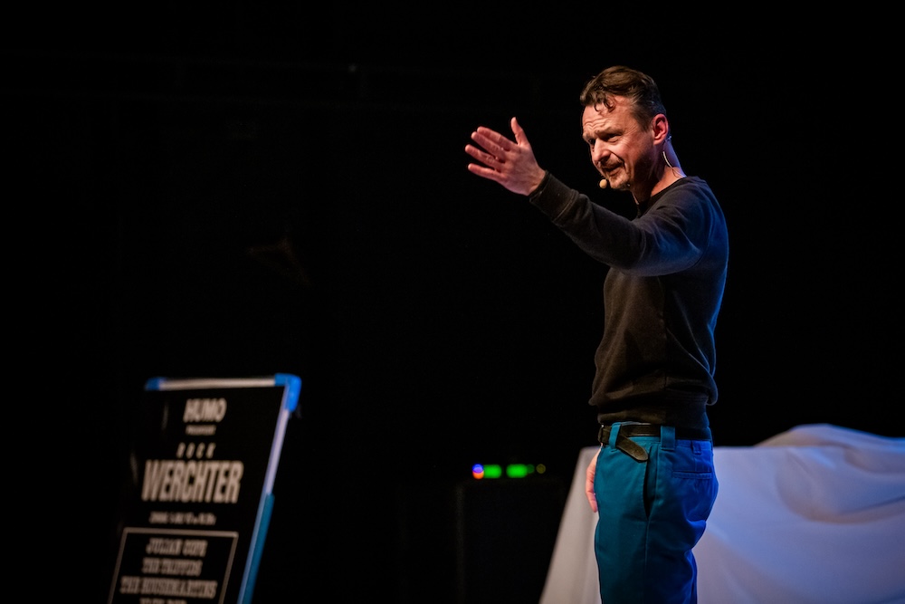 De Cock staat op een podium met een microfoonheadset en maakt een expressief gebaar met zijn hand, alsof hij het publiek toespreekt of een punt kracht bijzet. Op de achtergrond is een bord zichtbaar met 